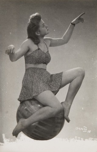 młoda kobieta, ujęcie całej postaci, siedzi na drewnianej kuli, zwrócona w prawo, ręce uniesione, ubrana w strój plażowy, tło neutralne, fotografia studyjna.