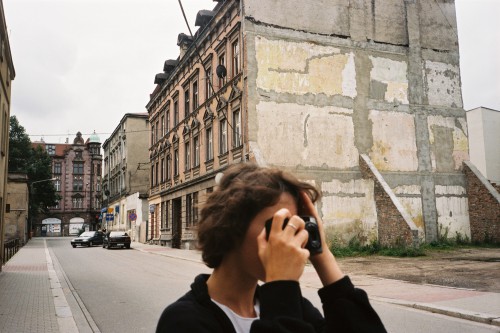 Kobieta stoi na ulicy, przy twarzy trzyma z aparat fotograficzny, gest fotografowania