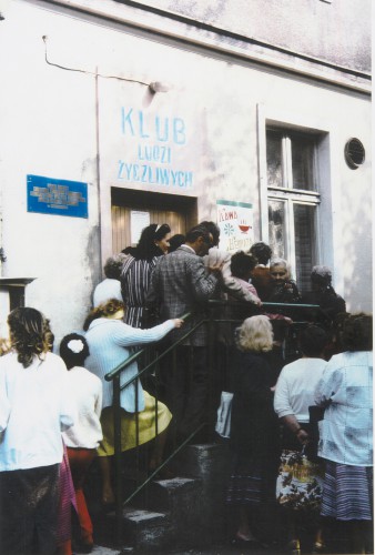 Grupa kobiet i mężczyzn w średnim i starszym wieku wchodzi po schodach prowadzących do drzwi zewnętrznych, nad drzwiami tablica z napisem: Klub ludzi życzliwych