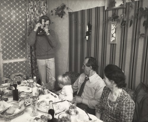 Kobieta, mężczyzna i dziecko siedzą przy stole, przed nimi stoi mężczyzna z aparatem fotograficznym wykonujący zdjęcie