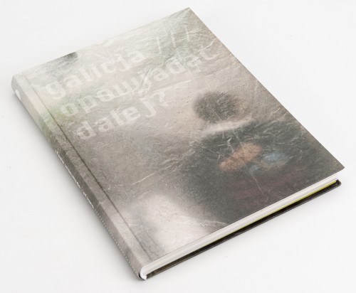 Leżąca książka, na okładce niewyraźne zdjęcie odwróconej tyłem postaci stojącej za szybą lub folią