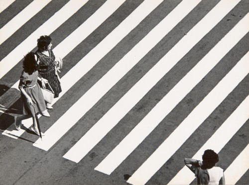 Fotografia czarno-biała, przedstawia widok z góry po przekątnej pasów ulicznych dla pieszych. Po lewej stronie widoczne z góry dwie kobiety ubrane w letnie sukienki. Pierwsza ma długie czarne włosy i trzyma parasolkę, druga, nieco starsza, ma krótkie czarne włosy i trzyma białą torebkę oraz przerzucony przez ramie jasny płaszcz. U dołu po prawej stronie widać odwróconą postać kobiecą przechodzącą przez pasy w przeciwnym kierunku