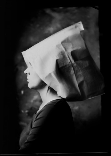 Fotografia czarno-biała, portret kobiety, lewy profil. Widoczna część twarzy, na głowie obszerne nakrycie przypominające papierową torbę lub arkusz papieru, kobieta ubrana w czarną bluzkę. Tło ciemne, mroczne