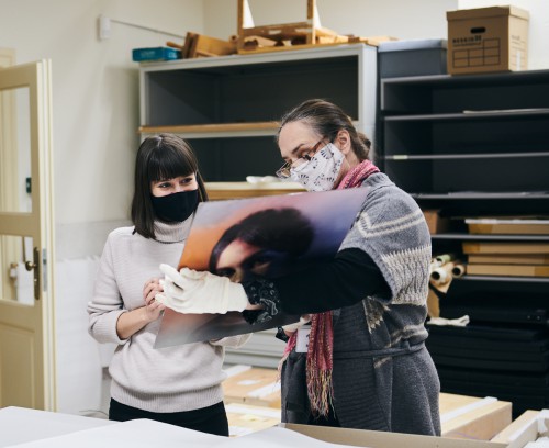 Dwie kobiety stoją na tle regału magazynowego, na twarzach maja maseczki. Jedna z nich trzyma w rękach kolorową odbitkę fotograficzną dużych rozmiarów i pokazuje ją drugiej. Na rękach ma białe rękawiczki do pracy konserwatora. Druga kobieta z uwagą patrzy na reprodukcję.