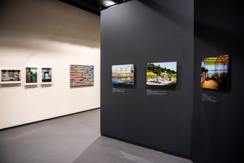 Na pierwszym planie pod skosem po prawej czarna ściana, na niej umieszczone w ramach w poziomym rzędzie cztery kolorowe fotografie,  po lewej w oddali biała ściana, na niej umieszczone w ramach w poziomym rzędzie trzy kolorowe fotografie.