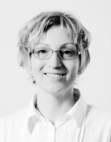 Fotografia czarno-biała, kobieta en fas w średnim wieku, włosy krótkie, pojedyńcze pasma opadające na twarz, okulary w owalnej oprawie, szeroko się uśmiecha, patrzy w obiektyw.