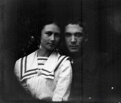 Fotografia czarno-biała, na czarnym tle para, przytuleni do siebie głowami.  Młoda kobieta ubrana w białą koszulę z dużym kołnierzem zakończonym dwoma ciemnymi pasami, meżczyzna ubrany w ciemny garnitur lub mundur. Patrzą w obiektyw, delikatne uśmiechy.