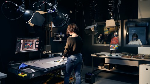 Na pierwszym planie kobieta stoi odwrócona przy blacie stołu. Na blacie wzornik kolorów, który widoczny jest też na monitorze komputera przed blatem. Wokół liczne lampy fotograficzne, aparaty i inne aparaty fotograficzne. Na blacie stojącym obok, po prawej stronie rozłożone w kupkach fotografie analogowe. Pomieszczenie zaciemnione, ściany częściowo oszklone. Za szybą po prawej stronie widoczna grupa zwiedzających przyglądających się pracy. 