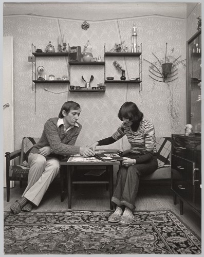 Scena rodzajowa, lata 80. XX wieku. Mieszkanie prywatne. Mężczyzna i kobieta siedzą pochyleni przy stole i oglądają album ze zdjęciami. Mężczyzna siedzi bokiem po lewej stronie, trzyma prawą ręką stronę albumu. Kobieta siedzi po prawej stronie, pochylona nad albumem, włosy półdługie ciemne częściowo zasłaniają jej twarz. Uśmiecha się. Obiema rękoma trzyma rozłożony na stole album. Nad parą wiszą 3 dwupoziomowe półki wypełnione bibelotami. Po prawej stronie kwietnik z pnącą się w dół ściany rośliną. Pod nogami rozłożony dywan. .
