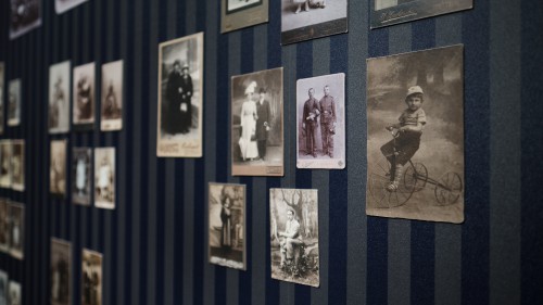 Pod lekkim ukosem ciemna ściana w podłużne ciemno-granatowe pasy. Na ścianie umieszczone bez ram fotografie atelierowe przedstawiające portrety z przełomu XIX i XX wieku.