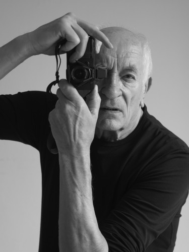 Portret starszego mężczyzny, ubrany w czarny golf z długimi rękawami, do prawego oka przystawia obiektyw aparatu fotograficznego, trzymany w obu rękach, aparat skierowany na wprost, w stronę obiektywu.