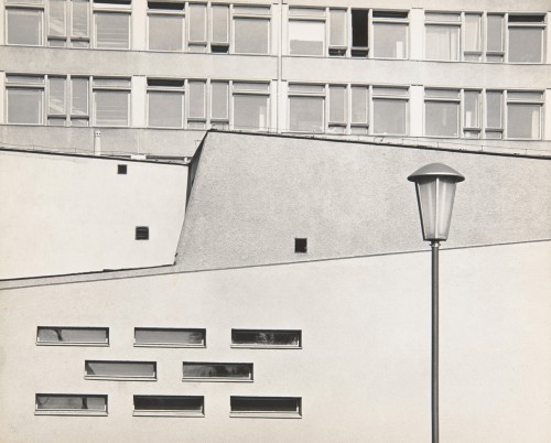 Nowoczesna architektura lata 70. XX wiek. Abstrakcyjny układ linii poziomych, pionowych i pośrodku - ukośnych. U góry dwie kondygnacje bloku z małymi kwadratowymi oknami. U dołu ozdobny układ 8 podłużnych poziomych okienek. Po prawej latarnia uliczna. 