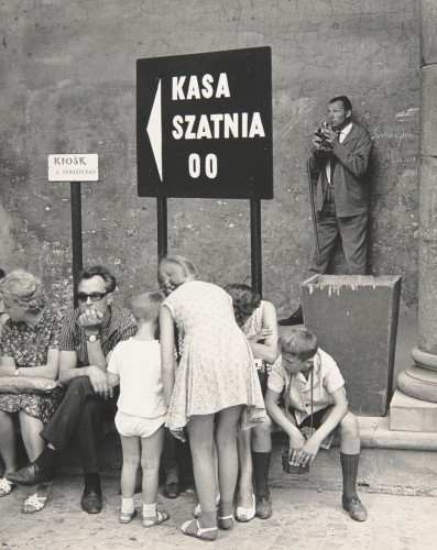 Na pierwszym planie 2 dzieci stoją tyłem, dziewczynka ok.10 lat, lekko pochylona, ubrana we wzorzystą sukienkę, sandały, po lewej chłopiec ok.5 lat w koszulce z krótkim rękawem, spodenkach, sandałach, nieco w głębi, frontem ku patrzącemu siedzą od lewej: kobieta, ok.40 lat, głowa w lewo, na kolanach torebka, ubrana we wzorzystą sukienkę, sandały, mężczyzna, ok.40 lat, w okularach, głowa wsparta na ręce, ubrany w koszulę w prążki, ciemne spodnie, półbuty, dalej dwie osoby widoczne częściowo. Po prawej chłopiec, ok.10 lat, głowa zwrócona w lewo, ręce opuszczone przed sobą, stopy szeroko rozstawione, ubrany w koszulę z krótkim rękawem, ciemne spodenki, podkolanówki, półbuty, na szyi pasek aparatu fot.,który przytrzymywany dłońmi,po prawej, na dalszym planie stojący mężczyzna,ok.30 lat,zwrocony 3/4 w lewo,ubrany w garnitur,białą koszulę,krawat,w dłoniach aparat fotograficzny,na przedramieniu prawej ręki laska. W centrum kompozycji kwadratowa tablica,przymocowana do stelaża,na ciemnym tle napis:KASA/SZATNIA/OO oraz strzałka, po prawej kosz na śmieci,po lewej prostokatna tabliczka: KIOSK/Z PAMIĄTKAMI,w tle fragment elewacji budynku:gładka powierzchnia pokryta tynkiem,częściowo spękanym, widoczne ślady uszkodzeń mechanicznych.