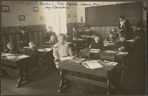 Dwa rzędy drewnianych ławek. W rzędzie po lewej chłopcy ubrani w mundurki. Po prawej dziewczynki w fartuszkach. Dzieci w wieku ok. 8 lat. Wykonują ćwiczenia plastyczne. Na ławkach rozłożone kolorowe papierki, nożyczki, klej. Na końcu sali pod ścianą z tablicą stoi nauczycielka. Ściany sali pokryte drewnianą boazerią, powyżej kolorowy pas (tapeta?). Na ścianach zawieszone obrazki dzieci. W przeciwległym rogu duże okno. 