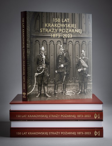 Książka, okładka z czarno-białym zdjęciem przedstawiającym trzech strażaków i złotym napisem, stoi na położonych na blacie czterech egzemplarzach tej samej pozycji