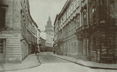 Ulica Bracka w Krakowie. Ujęcie w kierunku Rynku Głównego. Jezdnia i chodniki pokryte kocimi łbami. Widok obu pierzeji. W głębi górne kondygnacje wieży ratuszowej. Na pierwszym planie chodniki ulicy Franciszkańskiej pokryte płytami. 