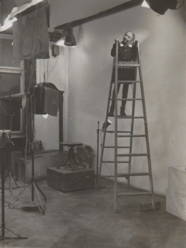 Studio fotograficzne Adama Karasia. Na drewnianej, wysokiej drabinie stoi dziewczynka w szkolnym chałacie. Lampy i wyposażenie studia fotograficznego: postumenty, taboret, tła fotograficzne zawieszone pod sufitem.
