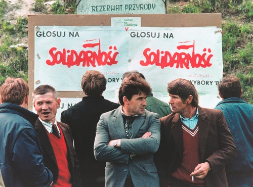 Grupa ośmiu mężczyzn, za nimi plakaty wyborcze z napisem: Głosuj na Solidarność. Lech Wałęsa Wybory 89.