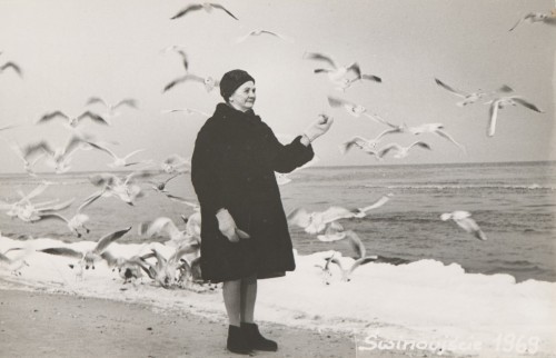 Portret starszej kobiety w czarnej czapce i czarnym płaszczu, stojącej na plaży. Wokół stado latających mew.