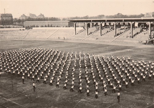 Widok na stadion z góry, ujęcie w perspektywie. Na murawie sportowcy z dzielnicy pomorskiej. Ustawieni równomiernie, przyklękają na jedno kolano, a ręce unoszą wysoko do góry.