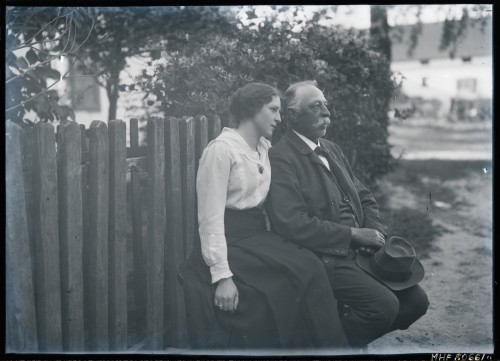 Na pierwszym planie młoda kobieta, ok. 20 lat, obejmująca lewą ręką mężczyznę, ok. 50 lat, o siwych włosach i sumiastych wąsach.