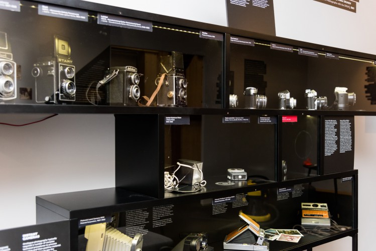 Wnętrze sali wystawowej, widok gabloty z trzema poziomami półek, na której wyeksponowane są stare modele aparatów fotograficznych. Pod każdym z modeli widoczny krótki opis.